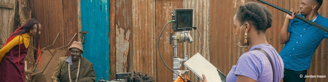 Ein Mann hält eine Tonangel und eine Frau hält ein Tablet und steht hinter einer Kamera. Vor der Kamera agieren zwei Darsteller*innen.