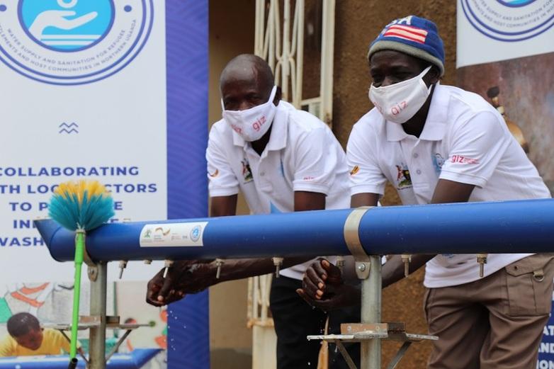 Two people wearing protective masks wash their hands at a group hand-washing station.