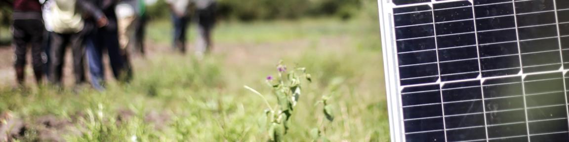 Ein Solarpanel steht auf einem Feld, im Hintergrund stehen mehrere Personen in einer Gruppe.