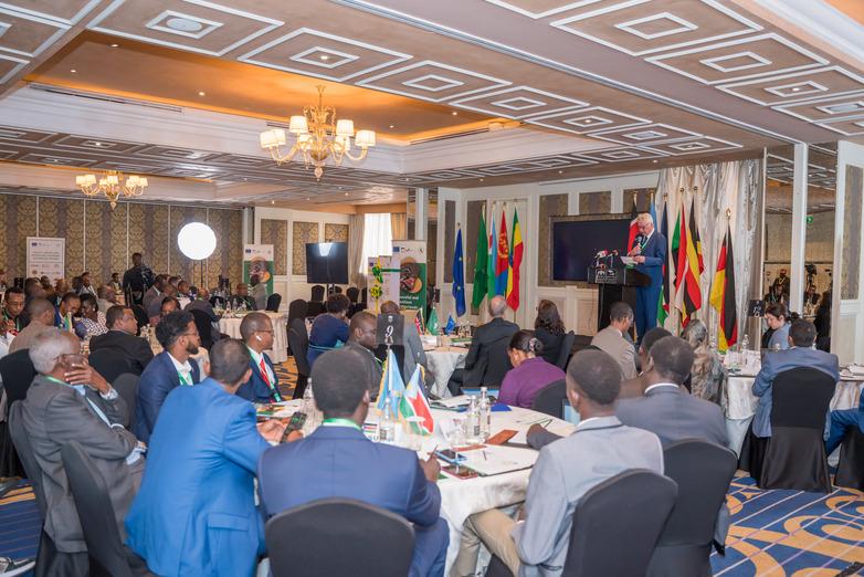 People are sitting at tables at an event. A man is speaking on stage.
