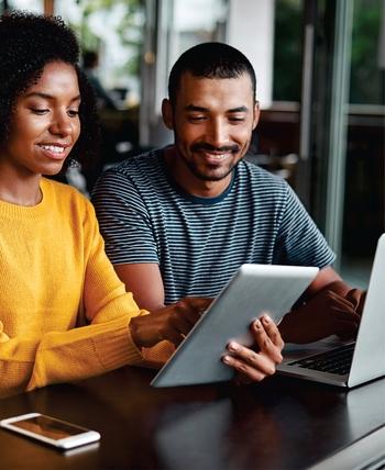 A young woman and a young man sit at a table. She is holding a tablet and he is using a laptop.