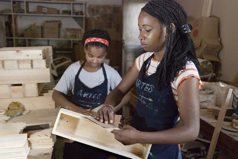 Two women trainees sand down small wooden crates in a workshop.
