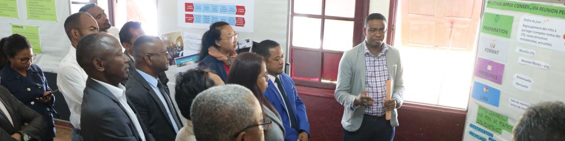 Des personnes sont debout devant un panneau de présentation, un homme parle.