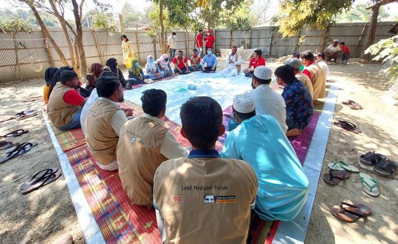 Participants sit in a circle on colorful mats during a meeting of the mediators' forum in a Rohingya camp, held outdoors and surrounded by trees and a fence.