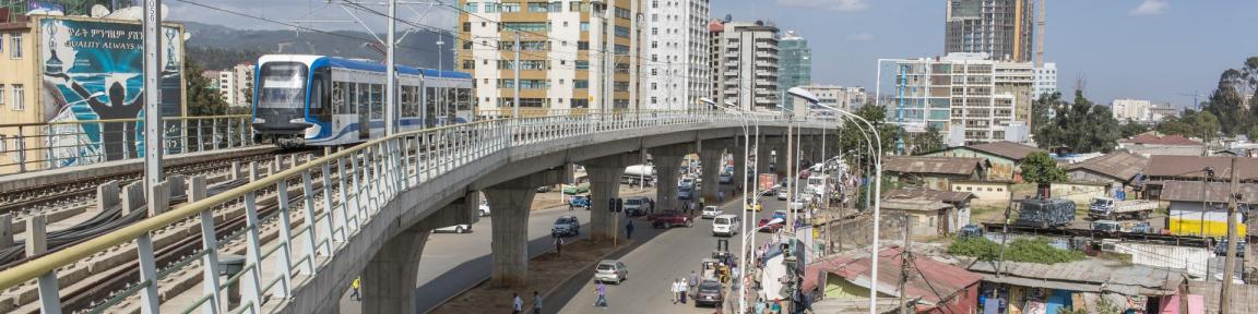 Ein blau-weißer Stadtbahnzug auf einer Hochbahnstrecke in einem belebten Stadtgebiet, umgeben von Hoch- und Flachbauten, bei klarem Himmel.