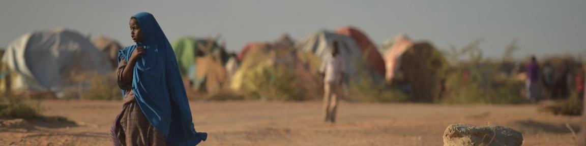 Eine Person mit einem blauen Schal geht in einem trockenen Gebiet in der Nähe von Zelten in einem Vertriebenenlager in Somalia.