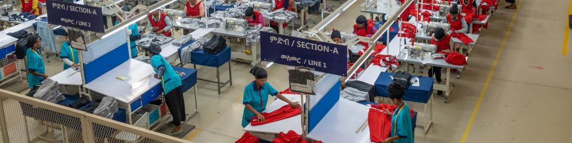 Blick von oben auf Westen tragende Arbeiter*innen, die in einer großen Bekleidungsfabrik reihenweise aufgestellte Nähmaschinen bedienen.