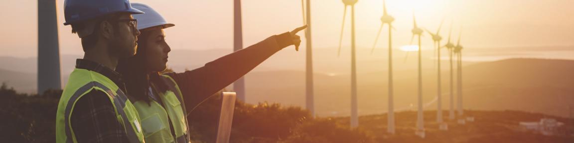 Zwei Arbeiter*innen stehen vor einer Reihe von Windkrafträdern und blicken in die Ferne.