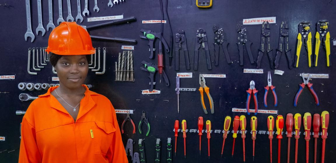 Eine Auszubildene mit orangenem Helm und Overall steht vor einer Wand mit Werkzeug.