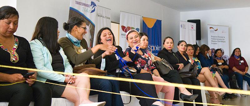 A group of women sit in a relaxed atmosphere in a room with posters in the background. The women are connected by colourful wool threads in their hands. 