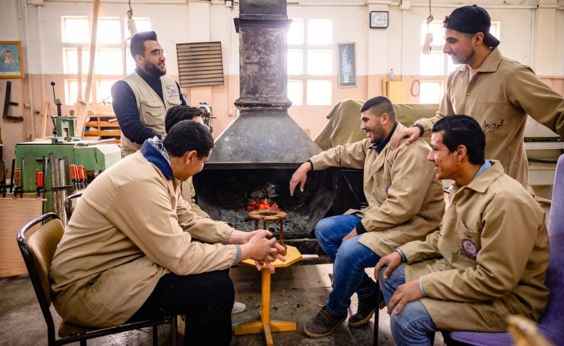 Young men in a vocational training workshop in Jordan gathered around a furnace, learning woodworking skills to enhance their employment opportunities.