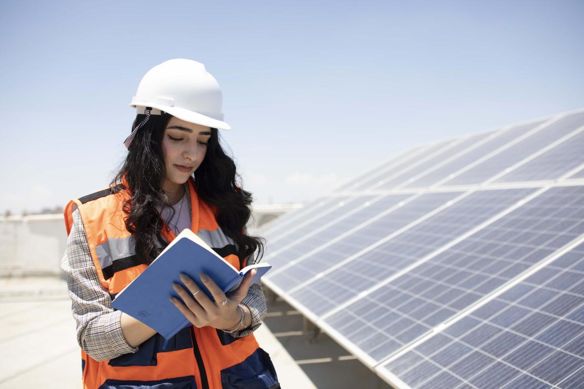 Eine Studentin mit Schutzhelm und Weste steht bei Solaranlagen und notiert etwas in einem Heft.