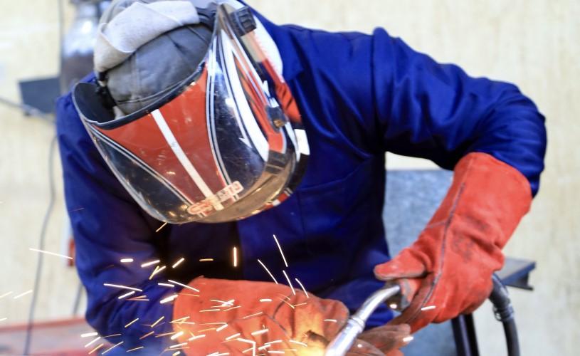 A worker in protective gear, including a welding helmet and gloves, welds metal in a workshop, producing bright sparks.