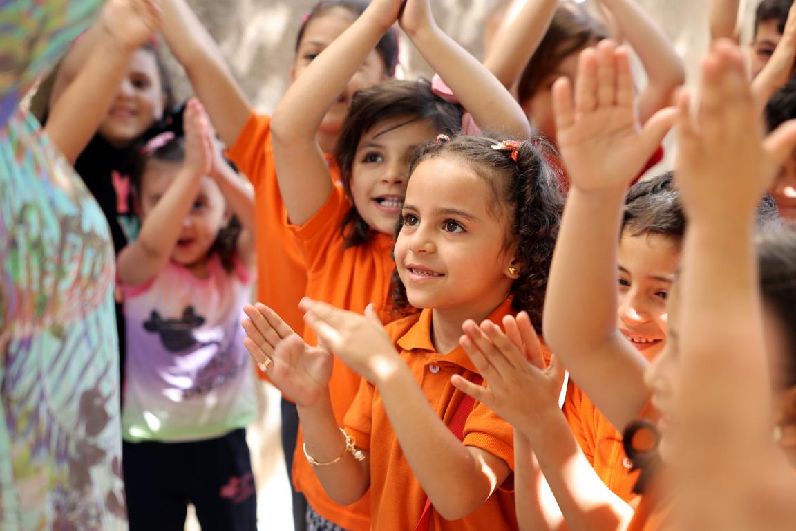 Children raise their hands and clap together during a group activity in a brightly lit classroom, participating in early childhood education in Jordan.