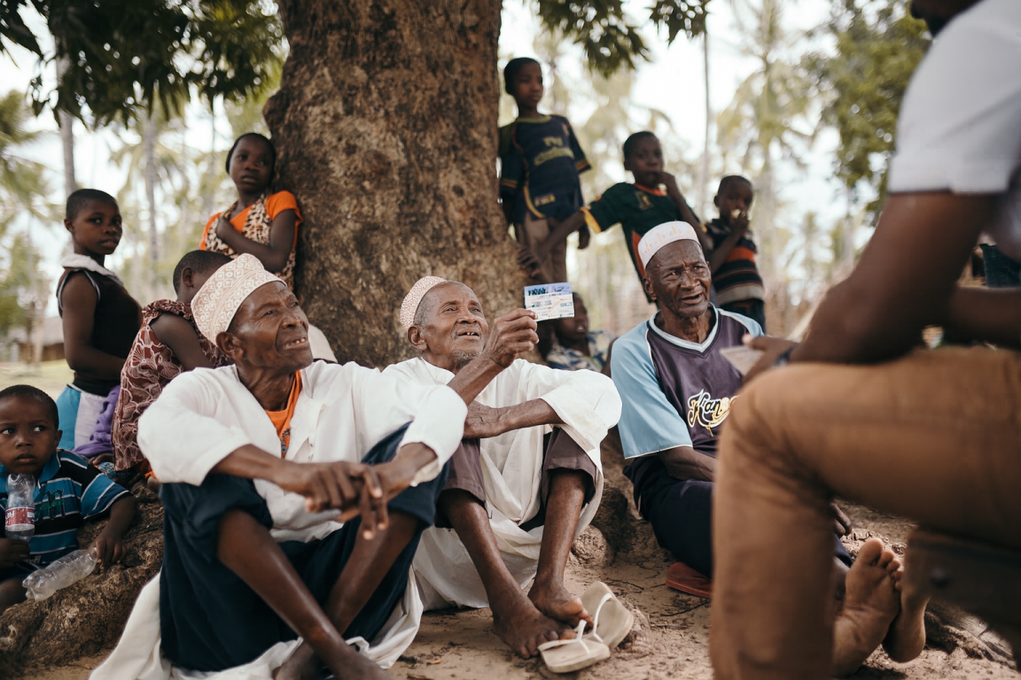 Several people sitting or standing next to a tree as they receive information about health insurance; one man is holding up a health insurance card. 