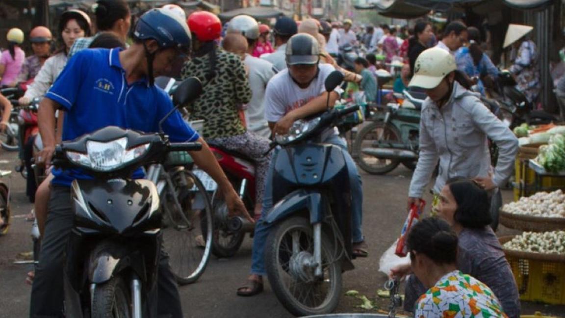 Geschäftiges Treiben auf einem Straßenmarkt in Vietnam: Menschen kaufen und verkaufen Waren und bewegen sich auf Motorrädern zwischen Marktständen mit frischen Produkten.