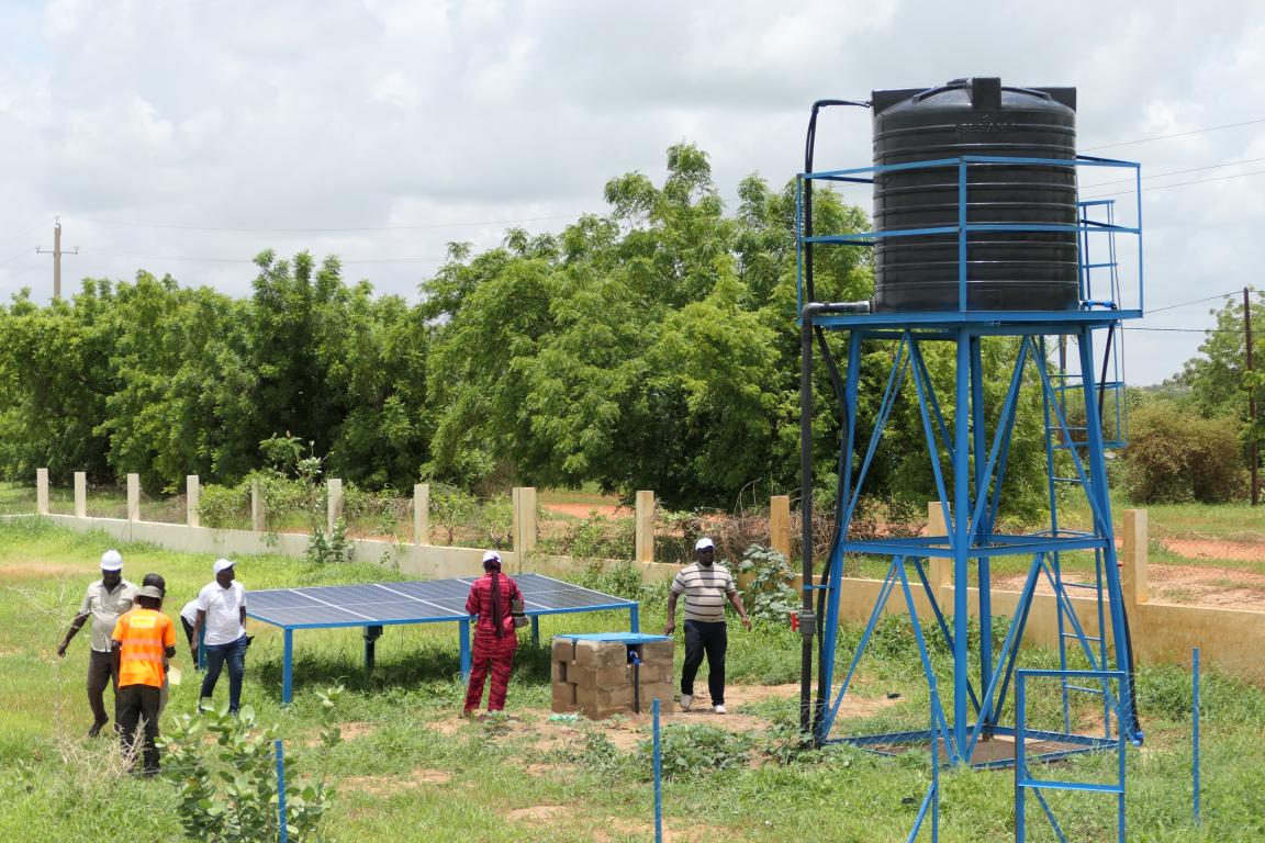 Menschen stehen auf einem Feld bei einem Wassertank, der mit einer Solaranlage betreiben wird.