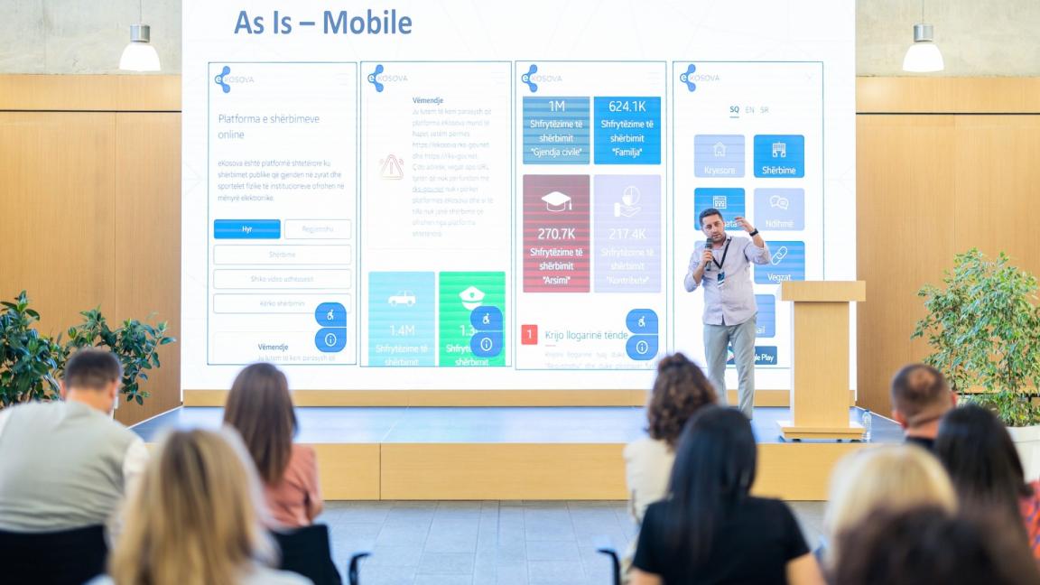 A presenter on stage pointing to a large screen displaying digital service statistics, during an event focused on e-governance and public service improvements in Kosovo.