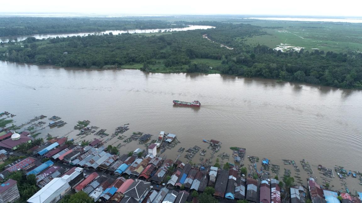 Aerial view of a river in Indonesia with a boat passing by and houses along the riverbank surrounded by lush greenery.