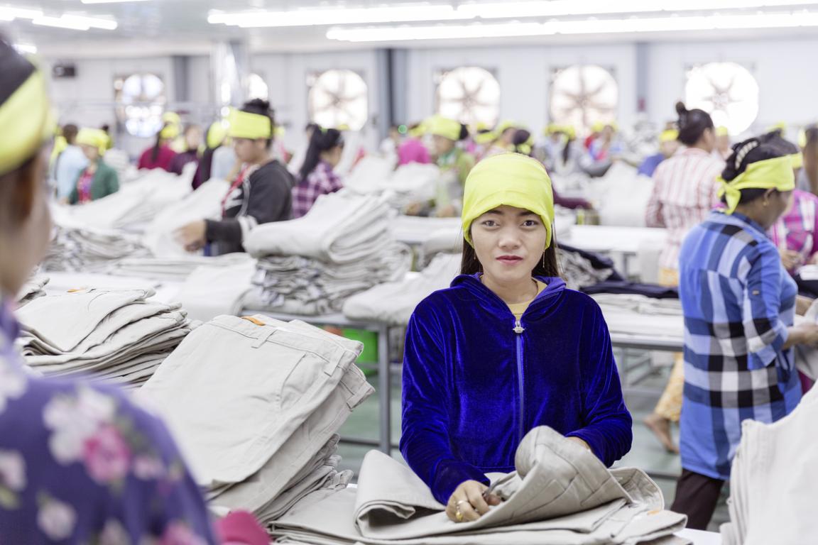 A woman working on a pair of trousers in a textile factory. Other workers are visible in the background.