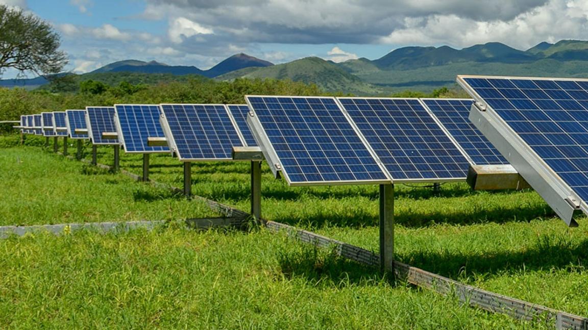 Rows of solar panels in a green field with mountains and clouds in the background.
