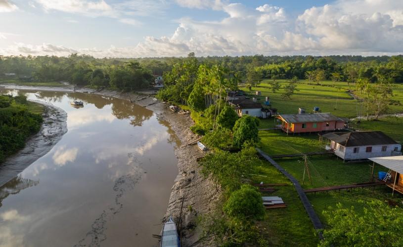 Aerial view of a riverside community in the Amazon region, with houses, trees, and boats along the riverbank.