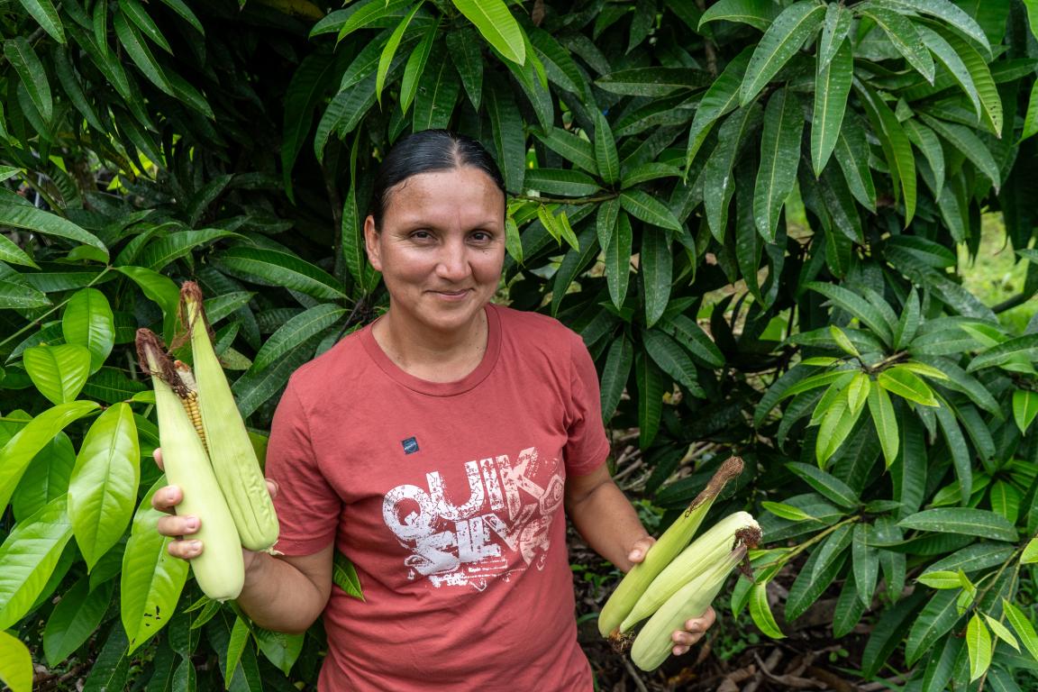 Una mujer está delante de un árbol y sostiene plátanos en la mano.