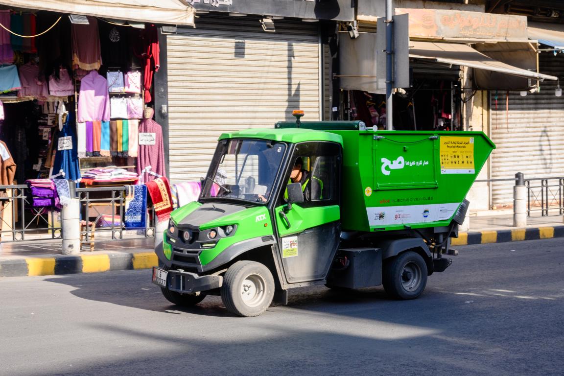 Ein kleines grünes elektrisches Müllfahrzeug fährt durch eine Straße mit Geschäften und geschlossenen Rollläden.