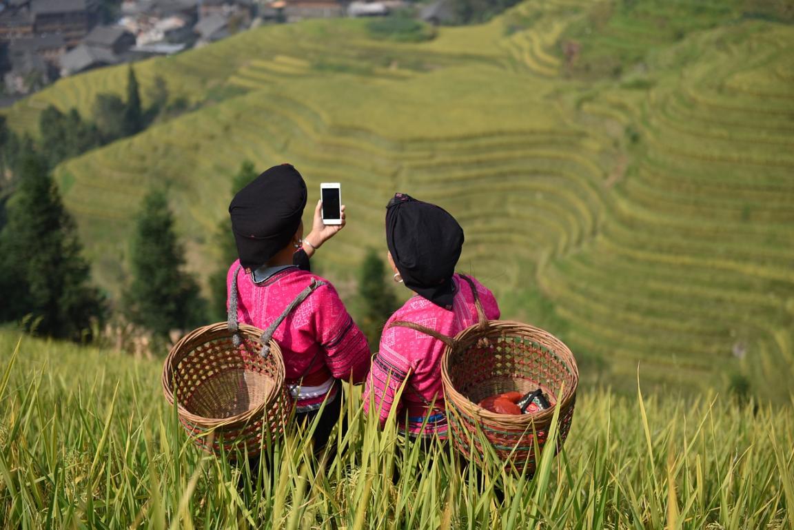 Zwei asiatische Frauen in traditioneller Kleidung stehen auf einem Feld und schauen auf ein Smartphone.