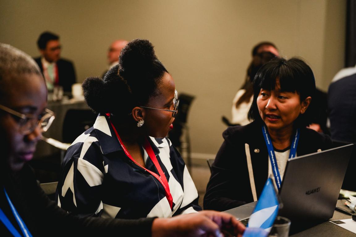 Several people sit at a table, two wearing conference badges and one with a laptop in front of them.