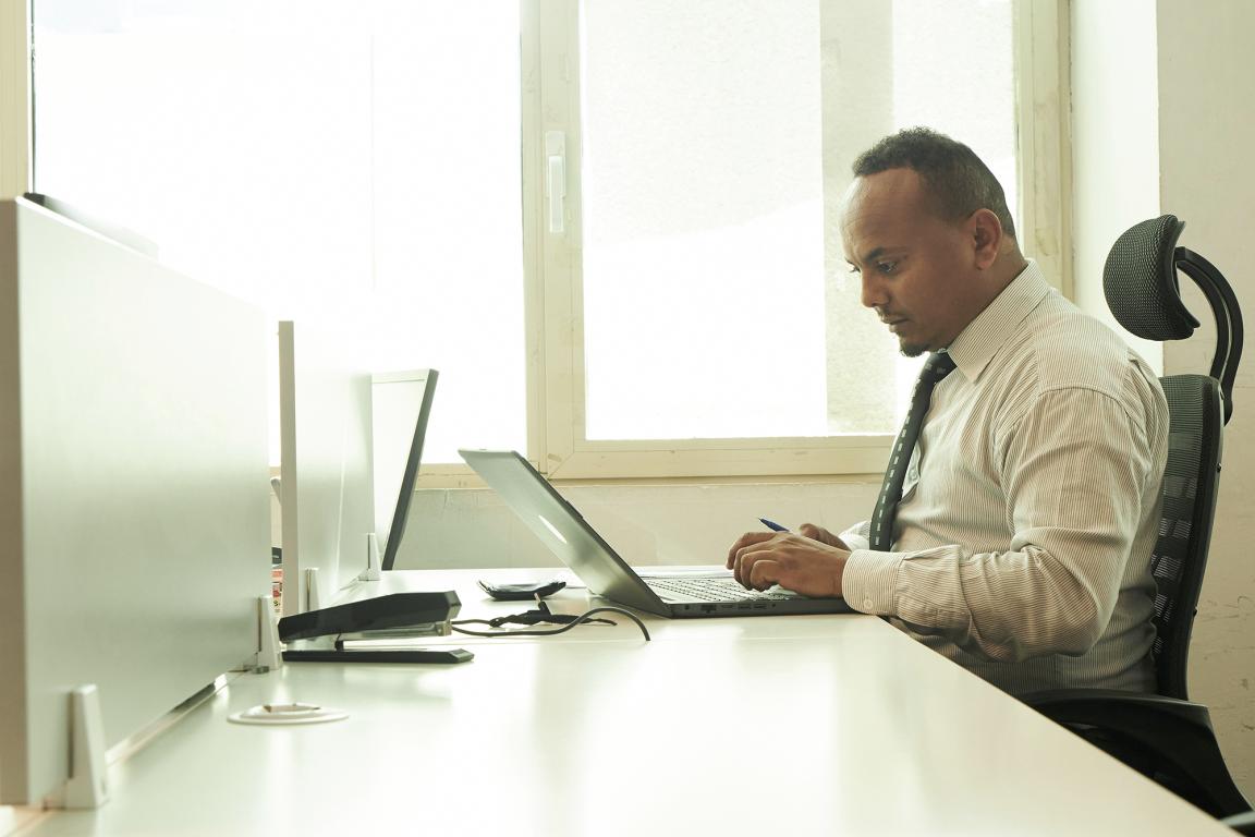 Man in shirt and tie sitting at a desk working on a laptop in a bright office with large windows.
