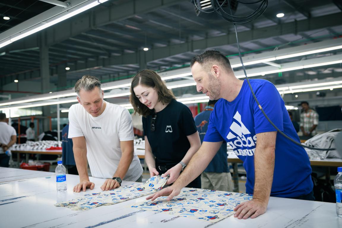 Trois personnes sont debout autour d'une table et examinent ensemble un morceau de tissu aux motifs colorés dans un environnement industriel lumineux.