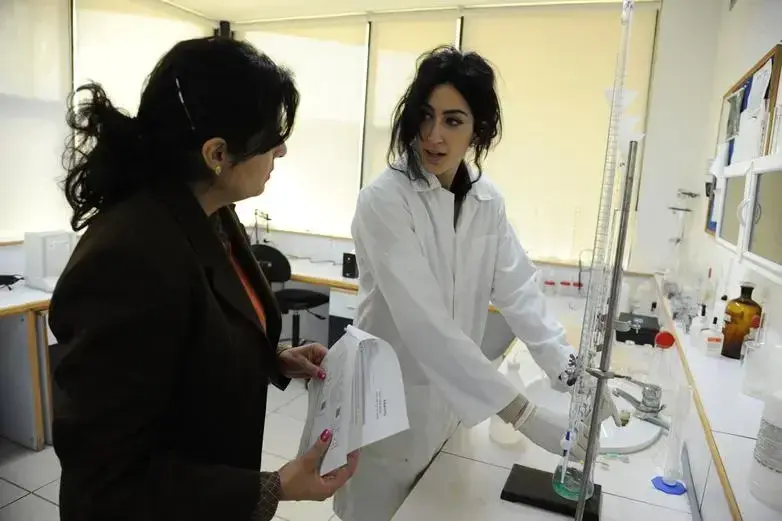Eine Frau in einem weißen Laborkittel zeigt einer anderen Frau an einem Arbeitsplatz ein chemisches Experiment.