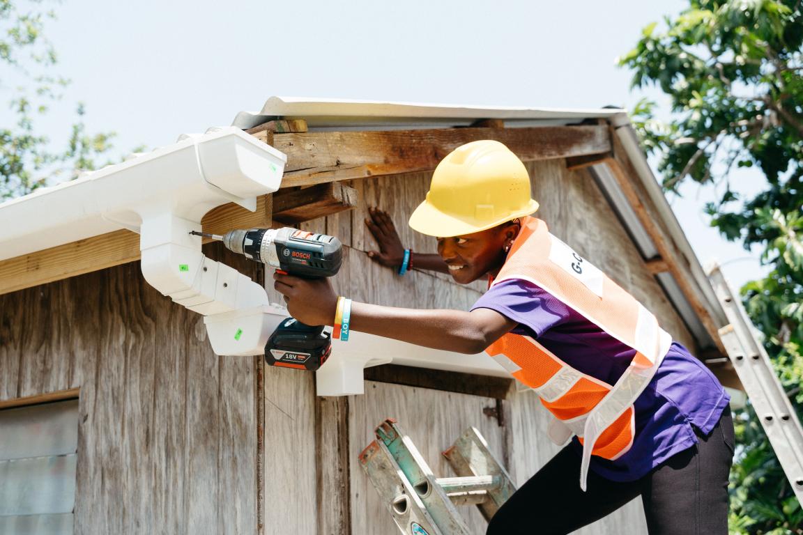 A young person in work clothes installing a gutter using a drill.