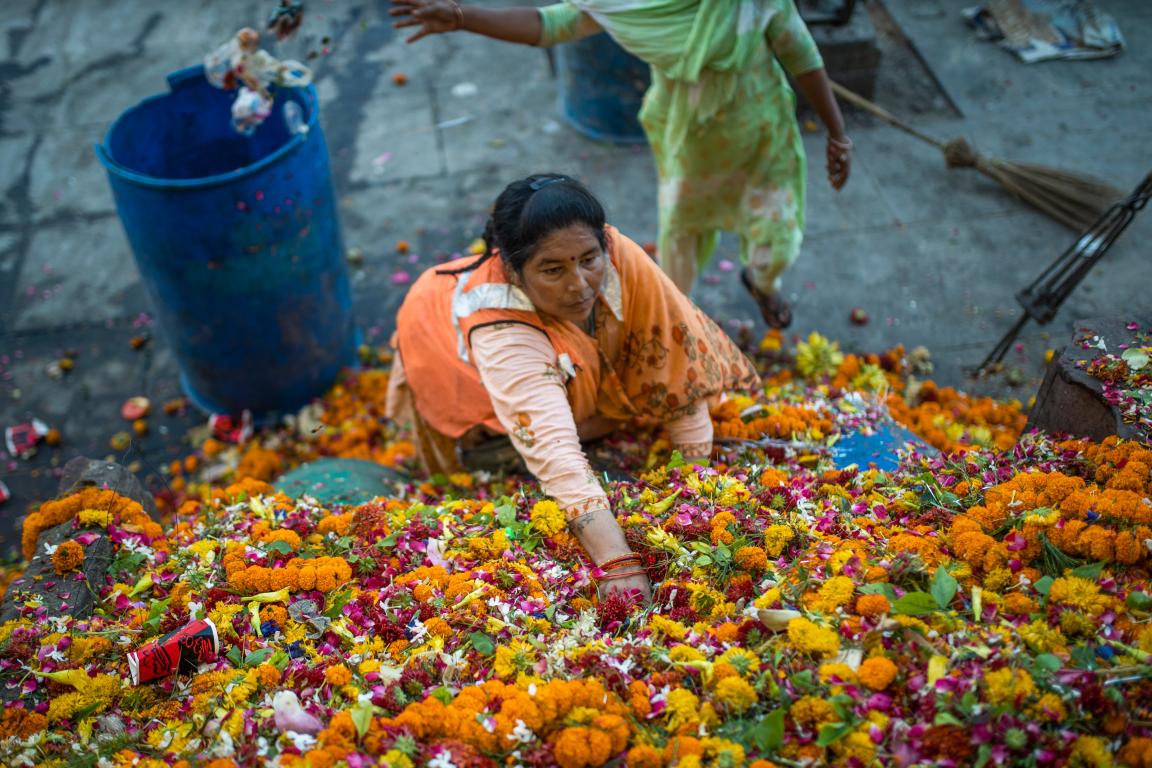 Eine Frau sammelt bunte Blumenabfälle aus einem Wasserbecken, umgeben von Blüten und weiteren Personen im Hintergrund.
