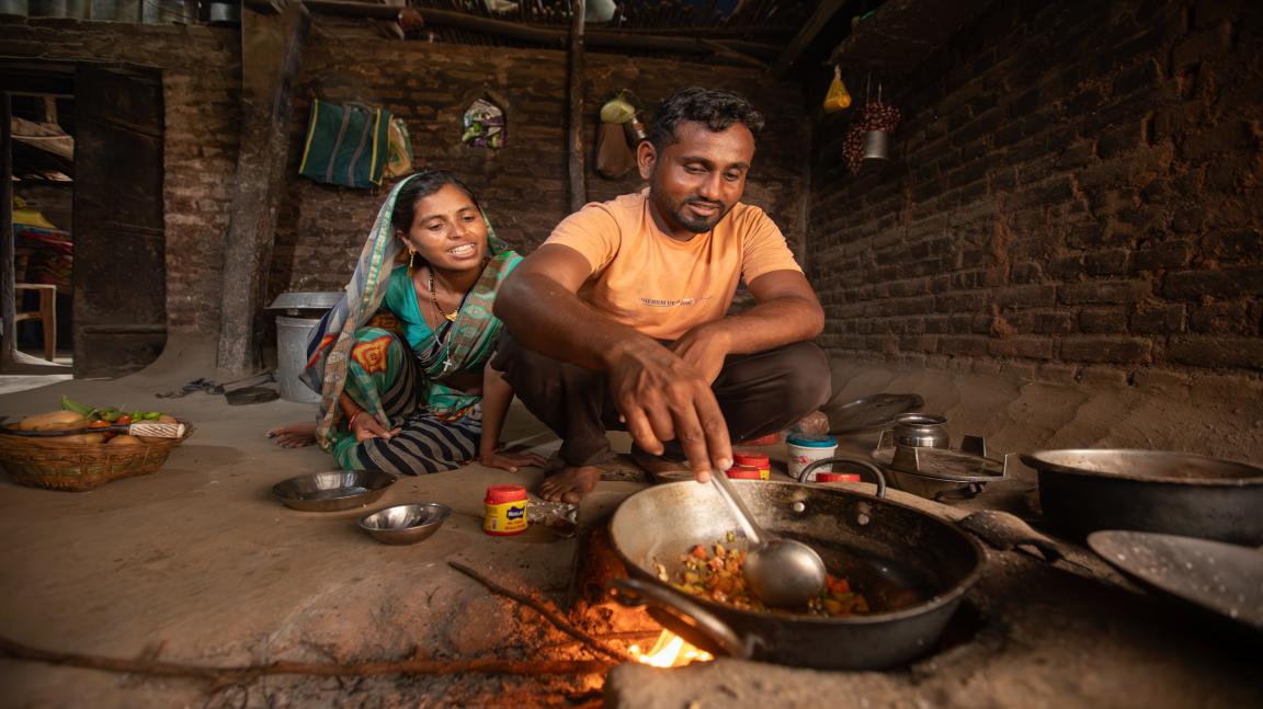 Two people sit on the floor in a simple room cooking in a pan over an open fire.