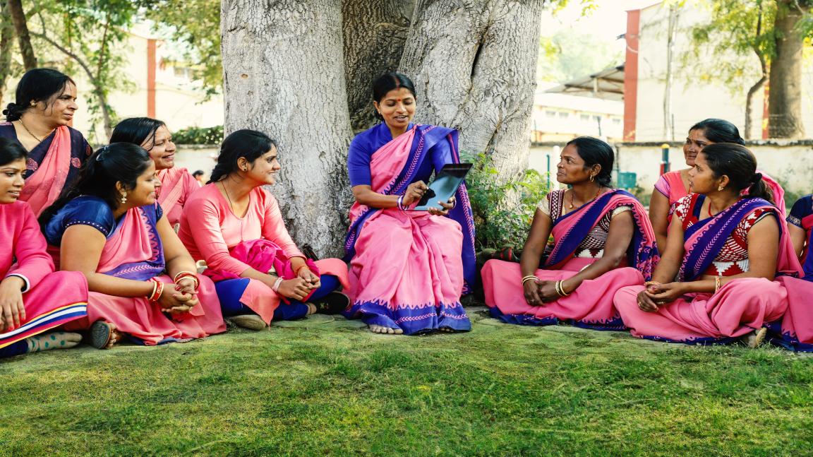 Group of women in traditional saris sitting in a circle on grass under a tree, one woman holding a tablet