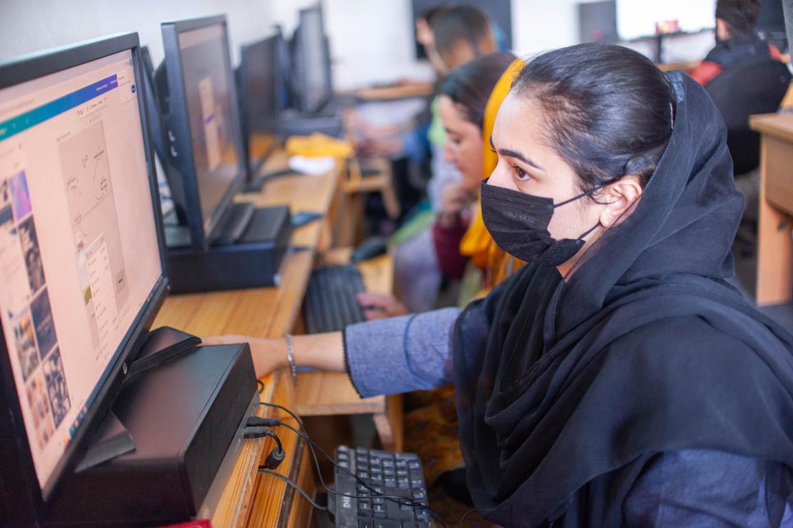 Woman wearing a headscarf working at a computer in a classroom with multiple people.