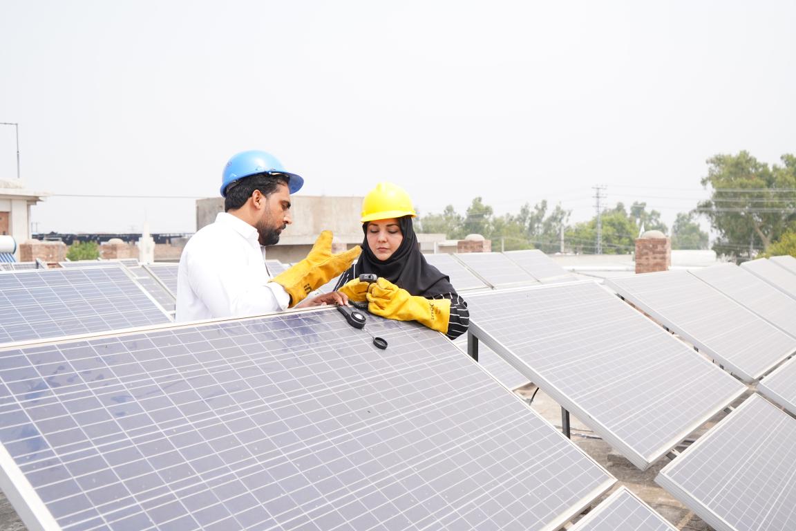 Two people wearing safety helmets and gloves stand on a roof with solar panels and talk to each other.