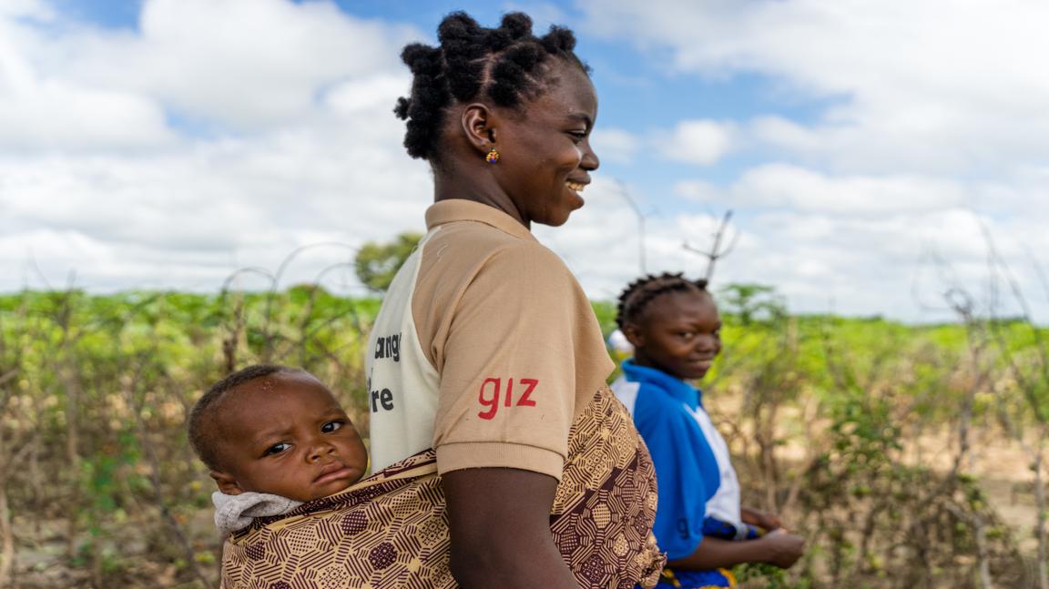 Une femme se tient devant un champ. Elle porte sur son dos un enfant en bas âge qui regarde l'appareil photo.