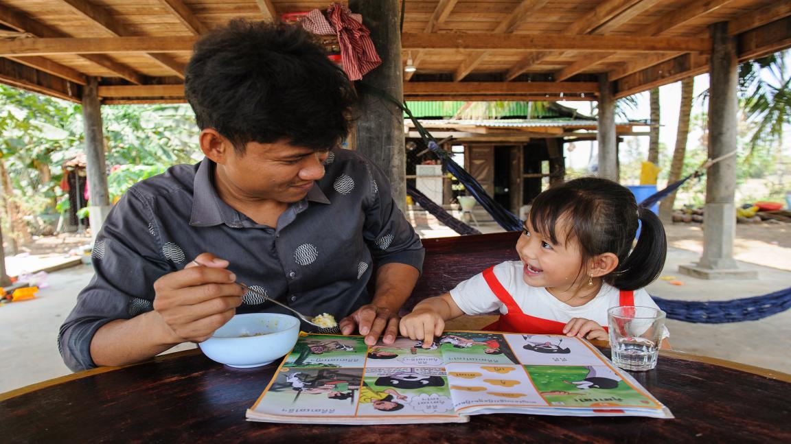 Un homme et un enfant sont assis à une table en bois sous un auvent extérieur. L'homme mange dans un bol, l'enfant montre une brochure colorée avec des images et des textes.