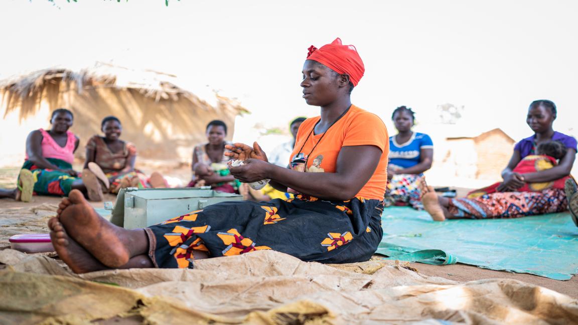 Une femme portant un foulard rouge et un t-shirt orange est assise par terre et tient quelque chose dans ses mains. À l'arrière-plan, plusieurs personnes sont assises par terre sous un toit de paille.