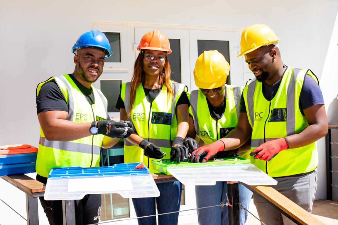 Four people wearing protective clothing, gloves, high-visibility vests, and safety helmets sort parts into boxes.