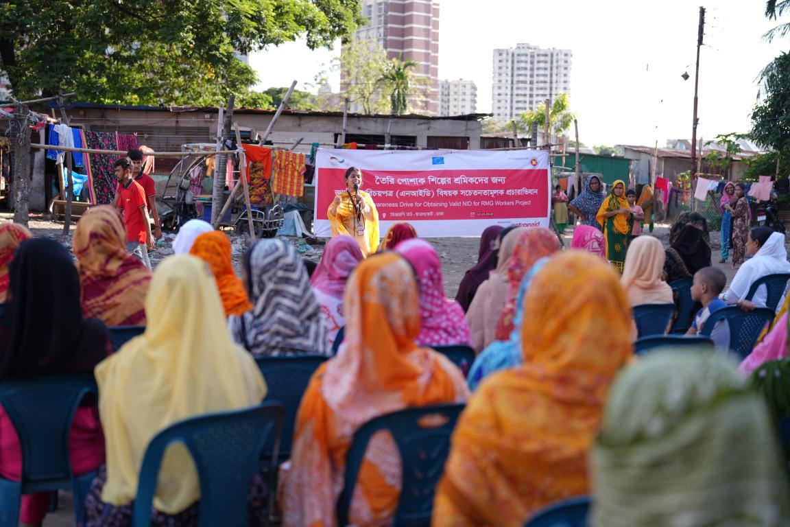 Gruppe von Frauen mit Kopftüchern sitzt auf Plastikstühlen im Freien vor einer Frau, die vor einem Banner spricht, im Hintergrund einfache Gebäude und Hochhäuser.