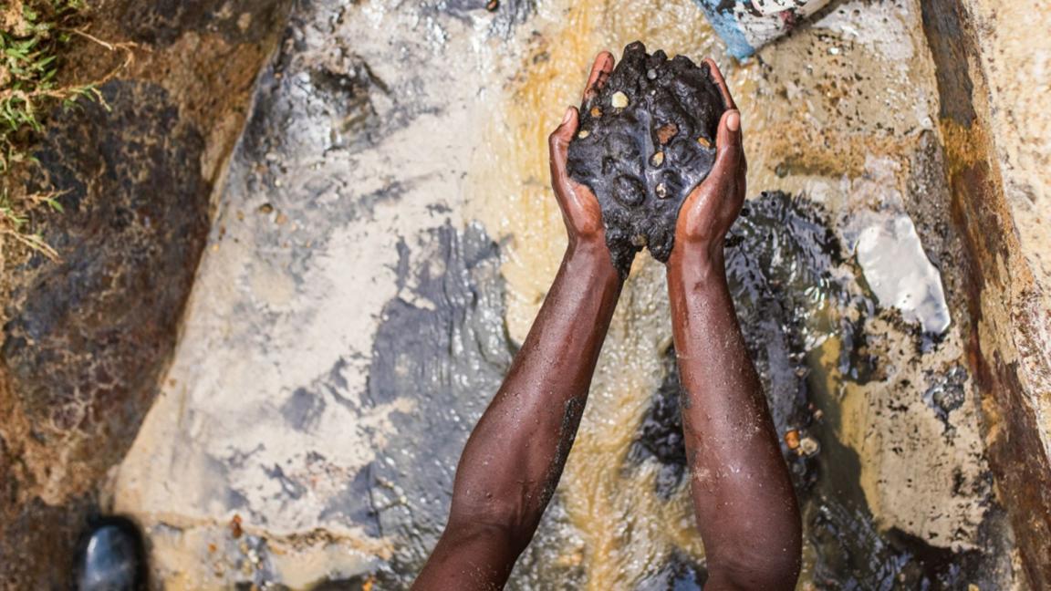 A miner holds a large, wet lump of freshly extracted ore in their hands above muddy water at a small-scale mining site.