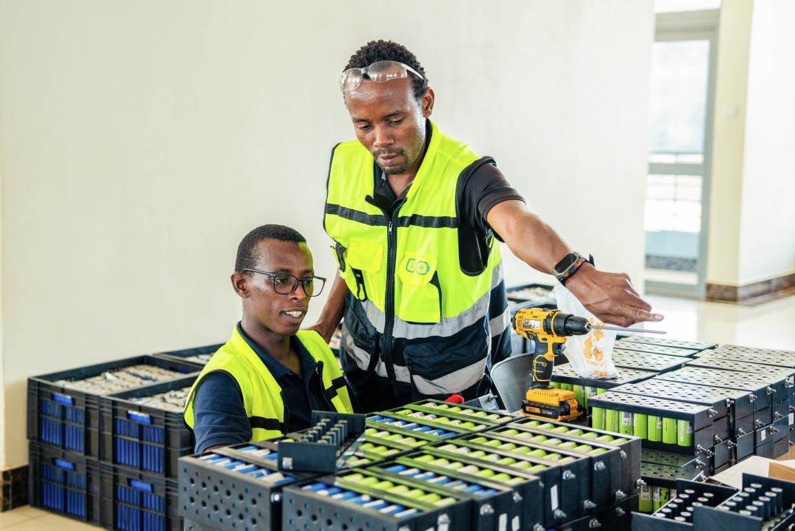 Two people in safety vests working at a table with many battery cells and a cordless drill