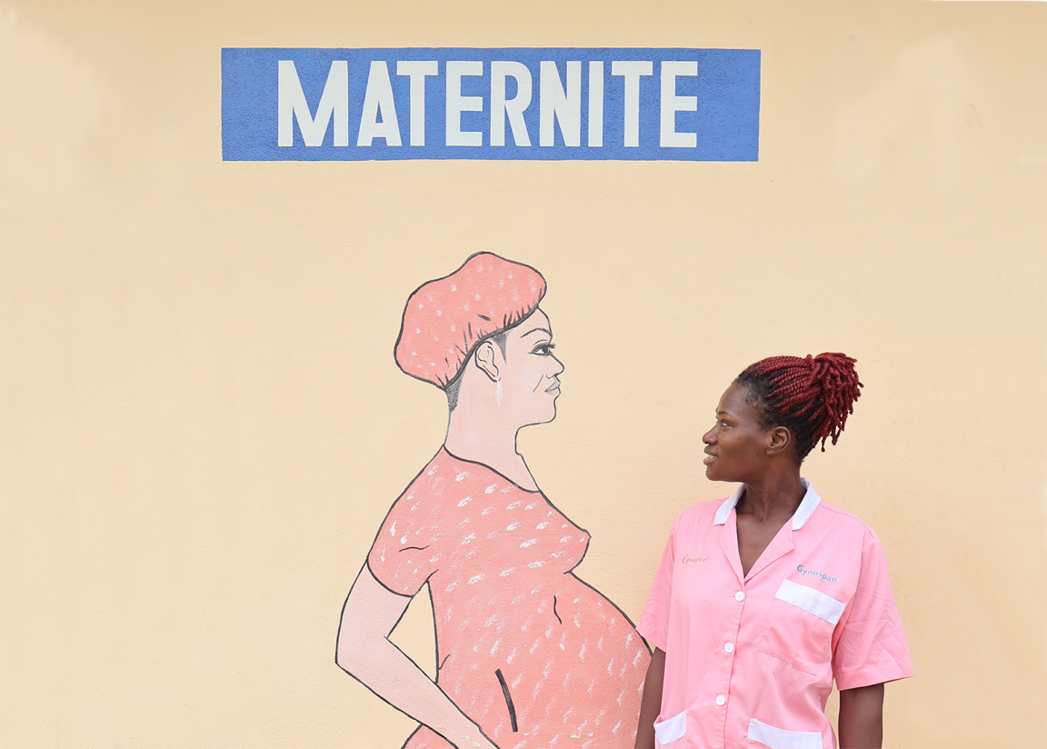 Woman in pink nursing uniform stands in front of a wall with a large drawing of a pregnant woman and the word 'MATERNITE'.