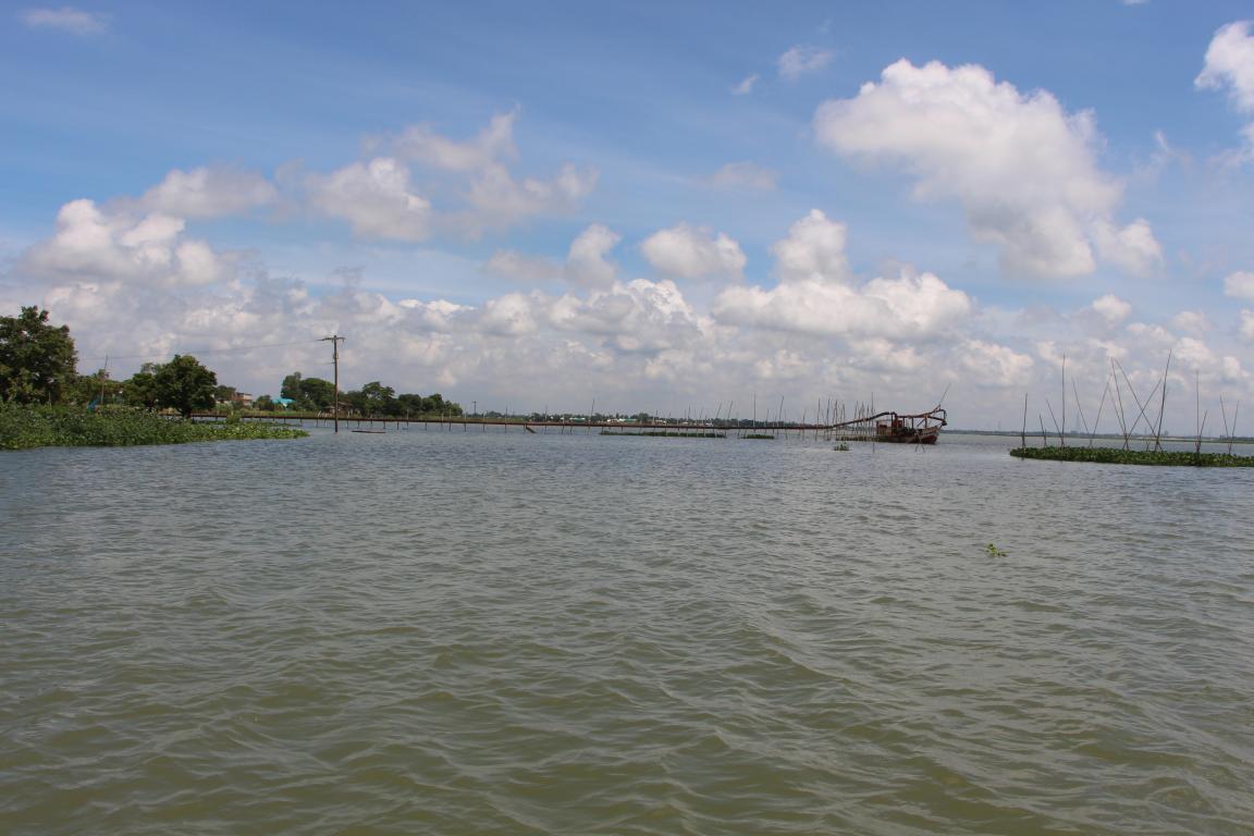 Fluss mit ruhigem Wasser, im Hintergrund Bäume, ein Steg und ein Boot unter bewölktem Himmel