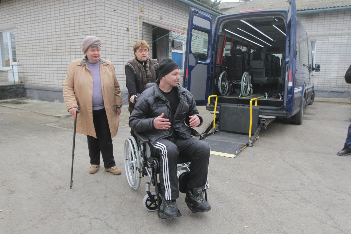 Чоловік у інвалідному візку з двома супроводжуючими біля автобуса з пандусом.