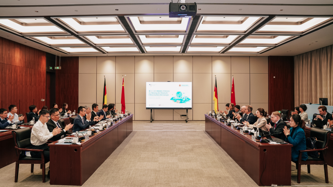 People sit at two rows of tables facing each other in a conference room and applaud; two German and two Chinese flags, as well as a screen, stand in the background.